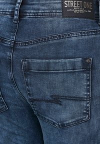 Blå denimjeans med en bakficka, som har ett subtilt slitet mönster och en läderetikett präglad med varumärket "STREET ONE."