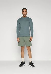 Fleecegenser i dempet blågrønn med logo, kombinert med lys grønne shorts og svarte treningssko.