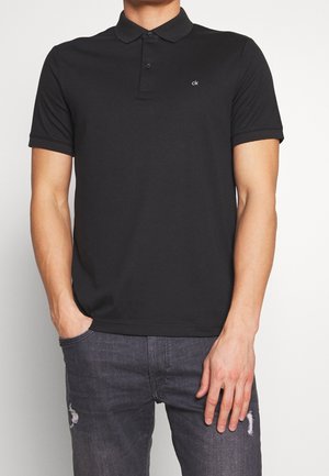 Mann trägt ein schwarzes kurzärmeliges Poloshirt mit kleinem "ck"-Logo auf der linken Brust und dunkelgraue, abgenutzte Jeans, steht mit einer Hand in der Tasche.