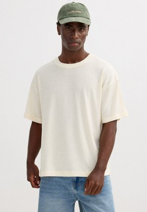 Joven con gorra verde y camisa de punto de manga corta en color blanco hueso, combinada con vaqueros azul claro, de pie frente a un fondo liso.