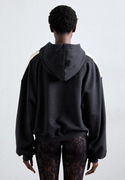 Black hooded sweatshirt with dropped shoulders, featuring cream side accents. Fabric appears soft with a loose fit. Worn with patterned leggings.