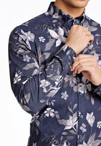 Camicia a bottoni con fantasia floreale su sfondo blu scuro. Presenta grandi motivi floreali grigi e bianchi, maniche lunghe e colletto.