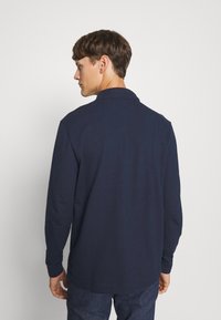 Marinblå långärmad polo med krage, med en slät yta och ribbade manschetter, visad bakifrån med synliga sömdetaljer.