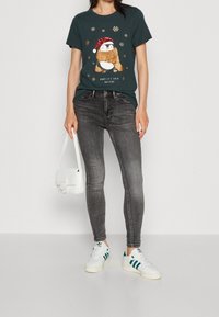 Camiseta de manga corta verde oscuro con un gráfico de pingüino, combinada con vaqueros gris ajustados y zapatillas blancas con acentos verdes. Bolso blanco.