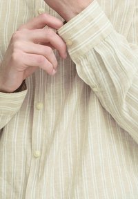 Beige stripete skjorte laget av lett stoff, med knapp mansjett og hvite vertikale striper med en svakt teksturert overflate.