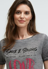 Gray, short-sleeve t-shirt featuring a round neckline and printed text "Soccx & Stories" and "LOVE" in pink with a textured finish.