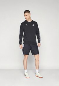 Felpa atletica nera con strisce bianche sulle maniche, abbinata a pantaloncini neri. Presenta i loghi di Adidas e Juventus, e scarpe da calcio bianche.