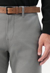 Grijze katoenen broek met een rechte snit, voorzien van een bruine leren riem en zichtbare stiksels. Het model draagt een zwarte trui.