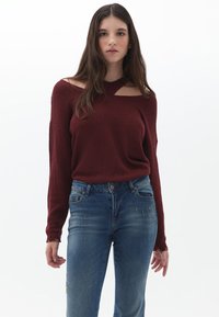 Burgunderfarbener Pullover mit offenem Schulterdesign, gestrickter Textur und langen Ärmeln, kombiniert mit blauen verwaschenen Jeans. Schlichter und moderner Stil.