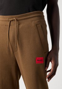 Dark-skinned person wearing brown sweatpants with a drawstring waist and red rectangular "HUGO" logo patch on the left thigh.