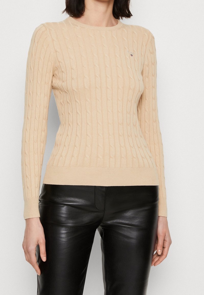 Jersey de punto color beige con mangas largas y dobladillo acanalado. La tela es suave y presenta un patrón texturizado. Se lleva con pantalones de cuero negro brillante.