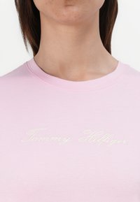 Artimas plano žmogaus, vilkinčio šviesiai rožinę marškinių, ant krūtinės išsiuvinėta kursyvine rašmena "Tommy Hilfiger" balta spalva.