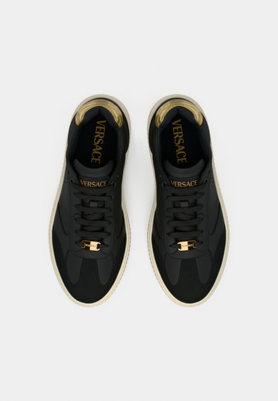 Czarne trampki o gładkiej fakturze, ze złotymi akcentami i białą podeszwą. Na języku widoczny jest wyraźny napis "VERSACE" oraz złote elementy ozdobne.