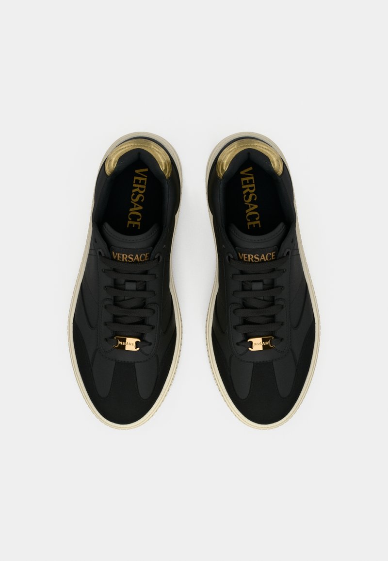Black sneakers with a smooth texture, gold accents, and a white sole. Features a prominent "VERSACE" logo on the tongue and gold hardware.