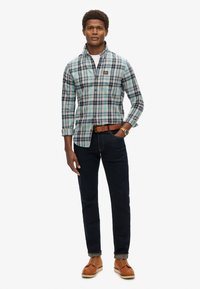 Camisa de cuadros en azul, verde y blanco, de manga larga, abotonada en el cuello. Combinada con jeans oscuros y zapatos marrones. Cinturón marrón incluido.