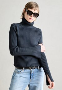 Maglione a collo alto nero a coste con maniche lunghe e fessure laterali, abbinato a jeans blu chiaro e una cintura nera. Tessuto testurizzato.