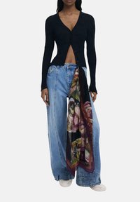 Femme portant un pull noir cropped à côtes, un jean bleu large avec un détail de foulard coloré, et des baskets blanches, se tenant devant un fond uni.