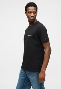 Μαύρο βαμβακερό μπλουζάκι με στρογγυλή λαιμόκοψη, εμπρόσθια τσέπη με κόκκινη, λευκή και μπλε ρίγα, συνδυασμένο με μπλε τζιν.