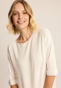 Jasnokremowa lekka bluzka z dekoltem w łódkę i rękawami trzyczwartkowymi, z delikatną strukturą prążkowaną i detalem w postaci guzika na mankietach.