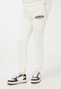 Jasno-beżowe dresy o dopasowanym kroju, z ściągaczami na końcach, z graficznym nadrukiem "Brooklyn Champions" w ciemnoczerwonym i czarnym kolorze na lewym udzie.