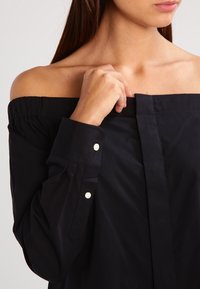 Svart off-shoulder tröja med långa ärmar och knäppta manschetter. Har en slät yta och en ren, minimalistisk design.