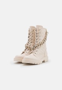 Stivali da combattimento in pelle beige con texture trapuntata, allacciatura frontale e dettaglio a catena dorata attorno alla caviglia. Suola spessa per una maggiore aderenza.