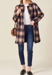 Károvaný kabát v námořnické modré a béžové barvě, zapnutý, s volným střihem. V kombinaci s modrými džíny a černými mokasíny, doplněný hnědou crossbody kabelkou.