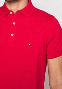 Polo rouge en mélange de coton avec une patte de boutonnage à deux boutons, manches courtes et un petit logo sur la poitrine gauche. Texture lisse, coupe classique.