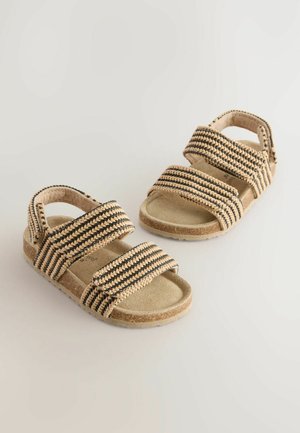 Beige sandaler med svarta vävda remmar, med två justerbara kardborrefästen och en texturerad sula för komfort. Platt sula.