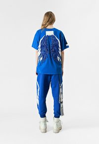 Camiseta de algodón azul con diseños de alas en blanco y azul en la parte posterior, combinada con pantalones de chándal azules a juego que presentan detalles en blanco y una etiqueta.