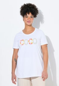 Camiseta blanca de manga corta con cuello redondo, que presenta letras de aplicación floral colorida formando la palabra "COCO" en el frente. Tejido de algodón suave.