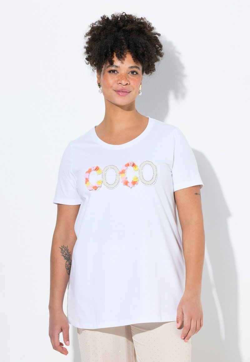 Camiseta blanca de manga corta con cuello redondo, que presenta letras de aplicación floral colorida formando la palabra "COCO" en el frente. Tejido de algodón suave.