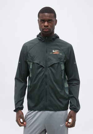 Atletski muškarac u tamnozelenoj vjetrovci s kapuljačom i Nike logom te sivim sportskim hlačama, stoji na jednobojnoj svijetloj pozadini.