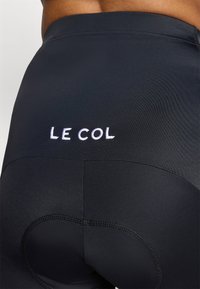 Svarte sykkelskjørt med reflekterende sømmer, med et brodert logo "LE COL" på baksiden. Laget av et glatt, elastisk stoff.