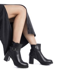 Botas de cuero negro hasta el tobillo con un tacón grueso, que cuentan con una cremallera lateral y un detalle de hebilla. La suela tiene un dibujo texturizado para mayor agarre.