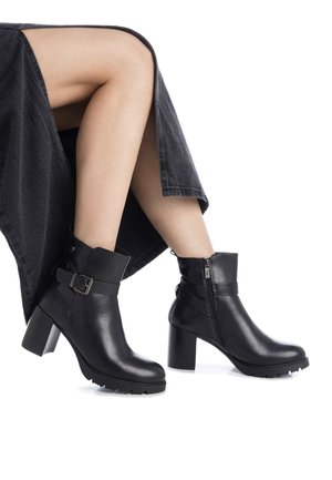 Botas de cuero negro hasta el tobillo con un tacón grueso, que cuentan con una cremallera lateral y un detalle de hebilla. La suela tiene un dibujo texturizado para mayor agarre.