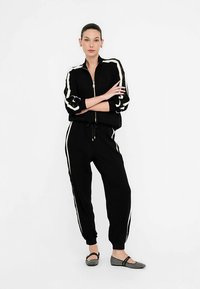 Sort træningsdragt med hvide sidestriber, lynlåstrøje, bukser med elastisk talje og mønstrede flade sko. Glat stof, afslappet pasform.