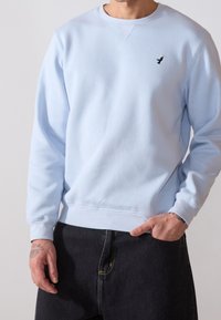 Felpa azzurra chiara realizzata in morbido tessuto con colletto rotondo, polsini a coste e un piccolo logo nero sul petto a sinistra. Abbinata a jeans scuri.