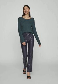 Suéter de malha de manga longa verde escuro combinado com calças justas de imitação de couro azul-marinho de cintura alta e sapatos de salto aberto pretos. Textura suave.