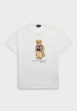 T-shirt en coton blanc avec un ours cartoon portant un manteau beige, une chemise bleue "POLO", un pantalon beige et des lunettes, avec du texte imprimé en dessous.
