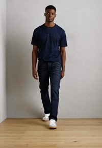Tricou bleu marin cu mâneci scurte, asortat cu blugi negri. Modelul poartă adidași albi. Design simplu, casual, cu o croială lejeră.