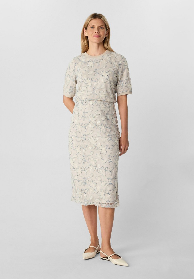 Femme debout portant une robe midi beige pâle brodée de fleurs à manches courtes et des ballerines beiges pointues, sur un fond uni.