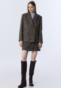 Blazer de cuadros marrón de doble botonadura con piedras decorativas, combinado con una falda corta a juego y botas de cuero marrón con tacón cuadrado alto.