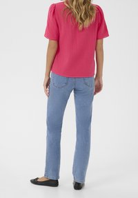 Rosa strukturierte Bluse mit kurzen Puffärmeln, kombiniert mit hellblauen geraden Jeans und schwarzen flachen Schuhen. Rückansicht des Outfits.