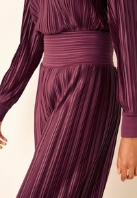 Robe plissée violet à manches longues et ceinture large et lisse, présentant une texture verticale et un tissu fluide.