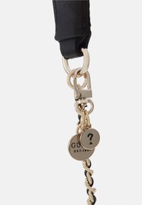 Gros plan sur une sangle noire avec une chaîne dorée et deux pendentifs en or, l'un gravé avec "GUESS EST. 1981" et l'autre avec un point d'interrogation.