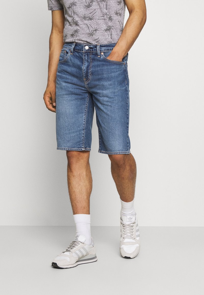Herrens blå denimshorts med rak skärning, fem fickor och synlig söm. Bärs med en gråmönstrad t-shirt och vita sneakers.