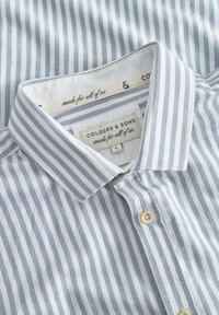 Camicia a righe azzurro chiaro e bianca con una texture morbida, dotata di colletto button-down e due bottoni davanti. Etichetta visibile all'interno.