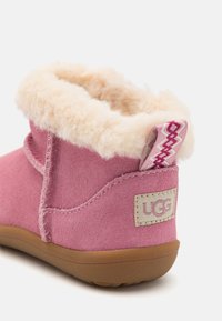 Botte en daim rose avec une douce doublure en shearling beige, dotée d'une languette décorative et d'une étiquette beige avec le logo de la marque. Semelle en caoutchouc.
