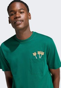 Zelené bavlnené tričko s vreckom a tromi malými grafikami žltých hviezdičiek. Krátke rukávy a dizajn s okrúhlym výstrihom.